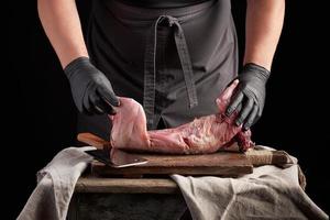 chef em luvas de látex pretas segura uma carcaça de coelho inteira em uma tábua marrom foto