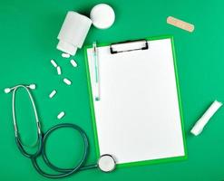 suporte de papel com lençóis brancos vazios, estetoscópio médico, comprimidos em um fundo verde foto