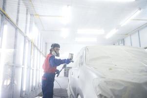 mecânico de automóveis asiático vestindo traje de proteção e respirador pinta pára-choques de carroceria na sala de pintura, pulverizando carros na sala de pintura foto