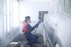 mecânico de automóveis asiático vestindo traje de proteção e respirador pinta pára-choques de carroceria na sala de pintura, pulverizando carros na sala de pintura foto
