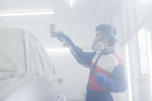 mecânico de automóveis asiático vestindo traje de proteção e respirador pinta pára-choques de carroceria na sala de pintura, pulverizando carros na sala de pintura foto
