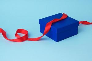 caixa de papelão quadrada azul para um presente e fita vermelha de seda torcida em um fundo azul foto