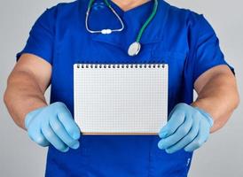 médico de uniforme azul e luvas de látex estéreis segurando o caderno em branco aberto foto