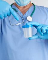 médico de uniforme azul e luvas de látex está segurando um recipiente de plástico vazio para coletar amostras de urina foto