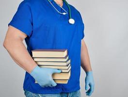 médico de uniforme azul e luvas de látex estéreis segura na mão uma pilha de livros sobre um fundo cinza foto