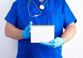 médico de uniforme azul e luvas de látex estéreis segurando o caderno em branco aberto foto