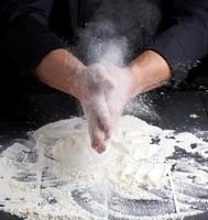 chef de uniforme preto derrama farinha de trigo branca de suas mãos em uma mesa de madeira foto