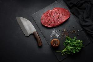 lombo de carne crua está sobre uma tábua e especiarias para cozinhar em uma mesa preta, vista superior foto