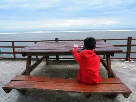 um menino de vermelho sentado em um banco de praia olhando para a bela praia e o céu foto