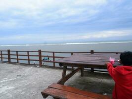 um menino de vermelho sentado em um banco de praia olhando para a bela praia e o céu foto