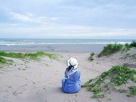 costas da mulher de chapéu na praia tropical que estava sentada na areia da praia. fundo de areia da praia e céu. montanha de areia. vista para a praia foto