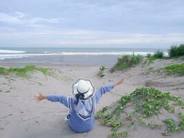 costas da mulher de chapéu na praia tropical que estava sentada na areia e olhando para o céu e o mar, enquanto abria os braços. mulheres felizes. vista para a praia foto