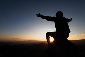 Silhuette jovem rezando na montanha, braços estendidos observando um belo nascer do sol dramático. foto