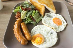 ovos fritos, salsicha, salada e torradas no prato e na mesa de madeira foto