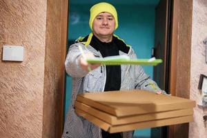 entregador com caixas de papelão de pizza. correio com chapéu de cor verde segurando uma prancheta em pé contra a porta da casa residencial foto