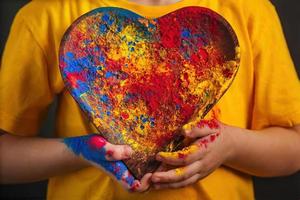 as mãos das crianças seguram uma placa de madeira em forma de coração na qual as cores brilhantes multicoloridas de holi. foto