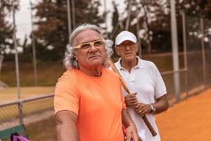 retrato de dois tenistas seniores vestidos com roupas esportivas relaxando no final do jogo em uma quadra de tênis de saibro foto