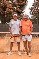 retrato de dois tenistas seniores vestidos com roupas esportivas relaxando no final do jogo em uma quadra de tênis de saibro foto