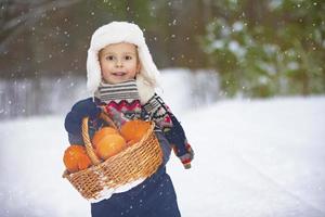 um menino feliz em roupas de inverno caminha pela floresta de inverno com uma cesta cheia de laranjas. criança com frutas no inverno. foto