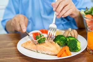 paciente senhora idosa asiática ou idosa comendo o pequeno-almoço de filé de salmão com alimentos vegetais saudáveis enquanto está sentado e com fome na cama no hospital. foto