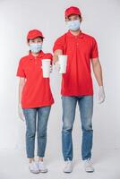 imagem de um jovem entregador feliz com gorro vermelho camiseta em branco uniforme máscara facial luvas em pé com copo de papel branco vazio isolado no estúdio de fundo cinza claro foto