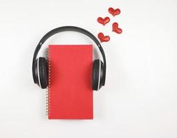 configuração plana de caderno vermelho coberto com fones de ouvido em fundo branco decorado com corações de glitter vermelho, copie o espaço. livro de áudio, podcast, diário .love, dia dos namorados. foto