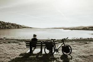 Ciclista caucasiano masculino pensativo sente-se no banco em turnê de bicicleta e desfrute do panorama do lago ao ar livre. memórias ativas do conceito de férias foto
