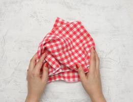 duas mãos femininas seguram um guardanapo de cozinha têxtil xadrez branco-vermelho amassado em uma mesa branca foto