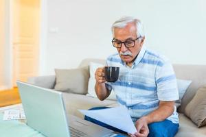 homem idoso de cabelos grisalhos focado senta-se no sofá lendo notificações bancárias calculando despesas domésticas, homem maduro moderno concentrado considera a papelada financeira, paga contas no laptop online foto