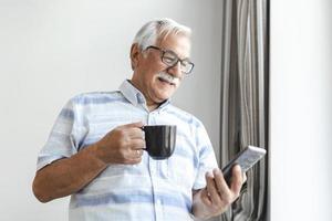 homem sênior usando smartphone. homem de cabelos grisalhos relaxando em casa lendo suas mensagens de texto em seu celular com um sorriso tranquilo. mensagens de texto masculinas sênior ou jogando um jogo online no smartphone em casa. foto