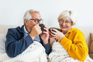 casal sênior se sentindo confortável e aquecido, sentado coberto com cobertor no sofá em casa. casal de idosos bebendo chá foto