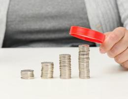 mulher senta às uma mesa e detém uma vermelho ampliação vidro sobre pilhas do branco moedas foto