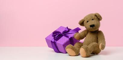 fofa Castanho Urso de pelúcia Urso segurando uma caixa embrulhado dentro papel e vermelho seda fita em branco mesa. prêmio e Parabéns, foto