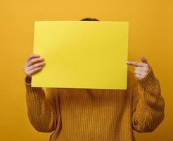 mulher com um suéter laranja segura uma folha de papel em branco sobre um fundo amarelo. lugar para uma inscrição, anúncio, informação foto