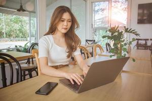 jovem inteligente ásia mulher freelance conectados trabalhando a partir de cafeteria com computador portátil computador e inteligente telefone. fêmea trabalhando remotamente às café fazer compras. conceito do trabalho em rede ou controlo remoto trabalhar. global o negócio rede. foto