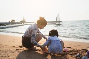 menina bonitinha asiática e sua mãe brincando ou fazendo castelo de areia ou cavando com areia na praia tropical. crianças com lindo mar, céu azul de areia. crianças felizes em férias à beira-mar na praia. foto