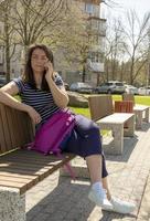 retrato de uma jovem morena atraente usando um smartphone, sentado em um banco de madeira em um parque e sorrindo. foto