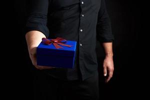adulto homem dentro uma Preto camisa detém uma azul quadrado caixa com uma vermelho arco amarrado em uma Sombrio fundo foto