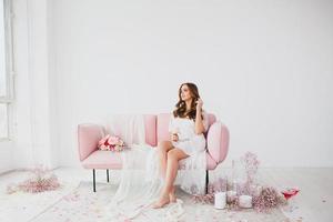 jovem noiva dentro branco lingerie posando em a rosa sofá dentro a estúdio foto