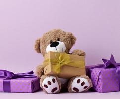 lindo ursinho marrom e caixa embrulhada em papel e fita de seda em um fundo roxo. prêmio e parabéns foto