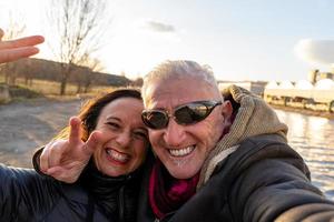 meio envelhecido casal vestindo inverno roupas levando uma selfie em a rio bancos foto
