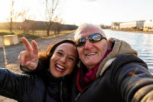 meio envelhecido casal vestindo inverno roupas levando uma selfie em a rio bancos foto