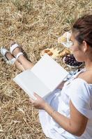jovem de vestido branco sentado no palheiro no campo colhido, lendo o livro em branco. maquete de livro foto