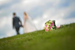 buquê de casamento na grama com um casal ao fundo foto