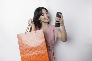 retrato ásia feliz lindo jovem mulher em pé animado segurando a conectados compras saco e dela Smartphone, estúdio tiro isolado em branco fundo foto