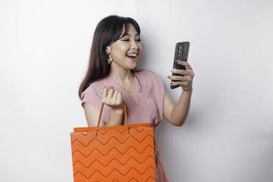 retrato ásia feliz lindo jovem mulher em pé animado segurando a conectados compras saco e dela Smartphone, estúdio tiro isolado em branco fundo foto