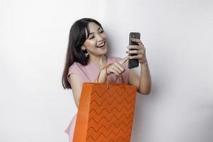 retrato ásia feliz lindo jovem mulher em pé animado segurando a conectados compras saco e dela Smartphone, estúdio tiro isolado em branco fundo foto