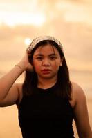 a ásia adolescente vestindo uma branco bandana e Preto camisa com uma plano expressão enquanto posando com dela cabelo em a de praia foto