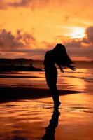 a ásia mulher dentro silhueta é fazendo uma muito lindo dança em a de praia com a ondas falhando foto