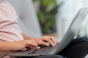 garotinha asiática da escola usa computador portátil sentado no sofá sozinho em casa. criança aprendendo lendo conteúdo de mídia social on-line, jogando lições de educação conversando com amigos. foto